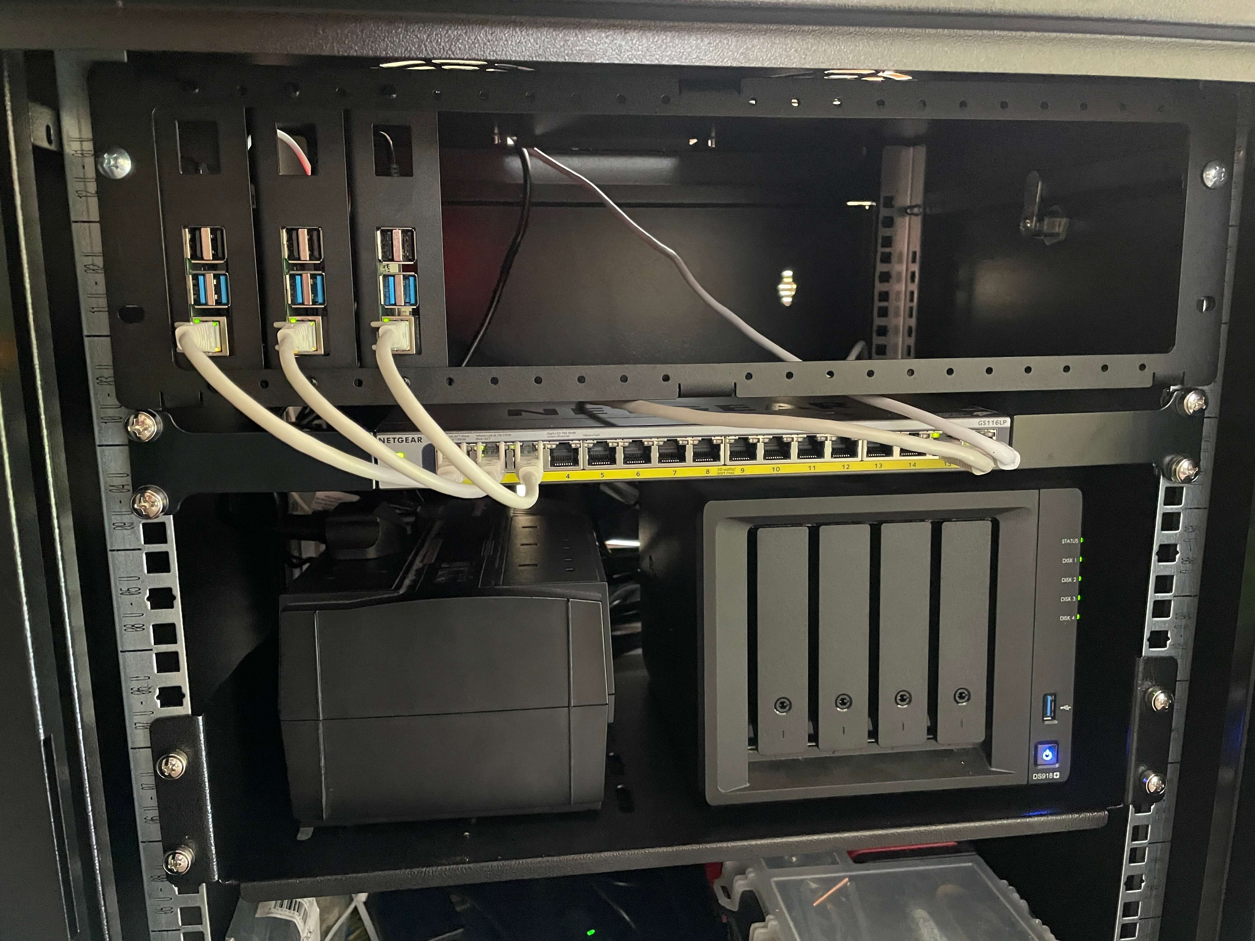 My home server rack from top to bottom: the 3 raspberry pis mounted in their mounting bracket, the PoE switch below them with patch cables running to each pi, and the shelf below that holding a UPS and a NAS